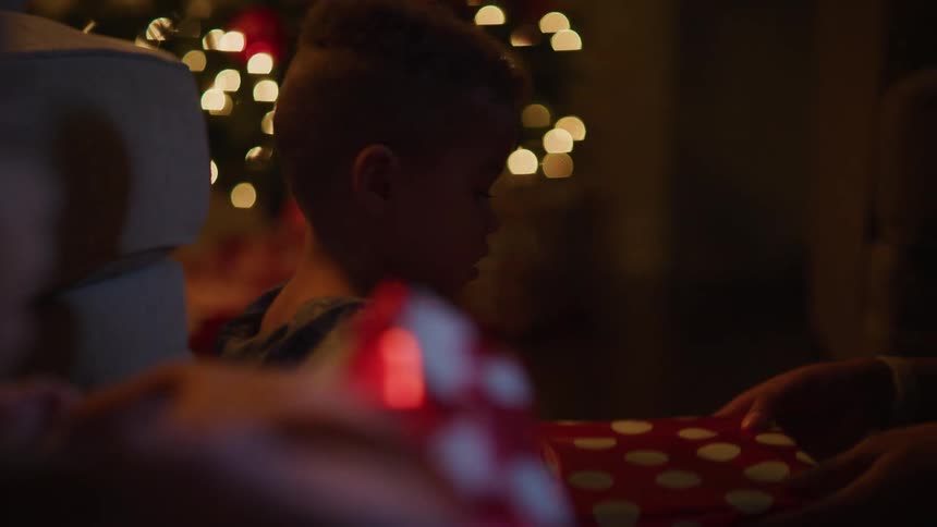 A young boy is handed a gift by an adult and begins to unwrap it while sitting indoors near a christmas tree
