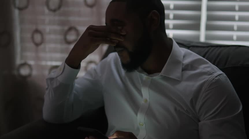 A man in a room sitting on a couch and looking at a smart phone with a window in the background