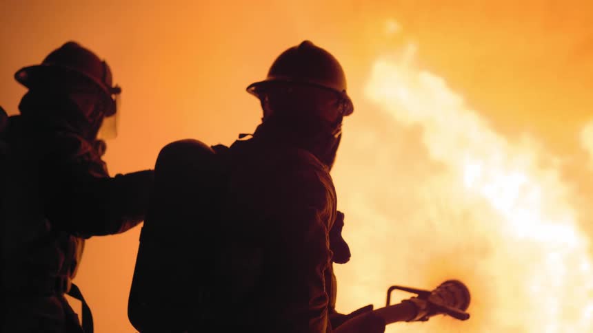 Silhouettes of firefighters battling huge flames with a hose