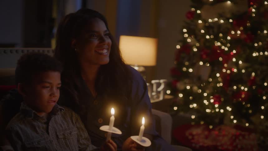 A mother laughs and puts her arm around her son as they sit on the couch next to a christmas tree and hold candles