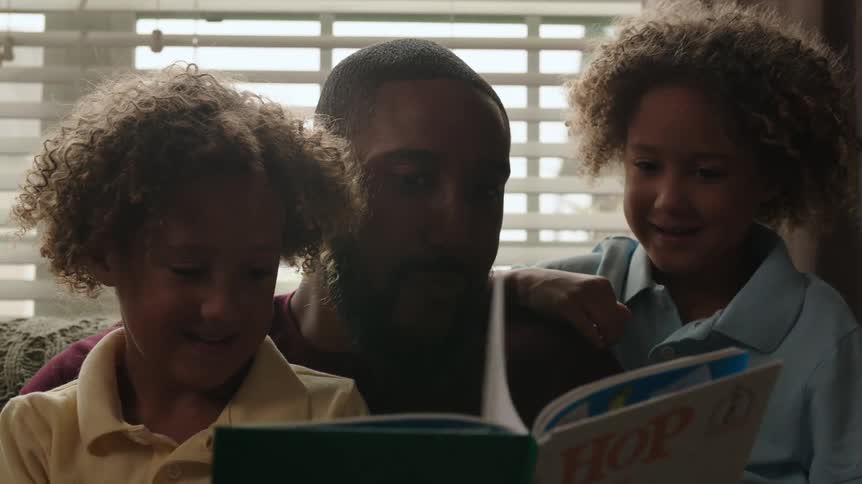 A man and two children in a room sitting on a couch reading a book with a window in the background