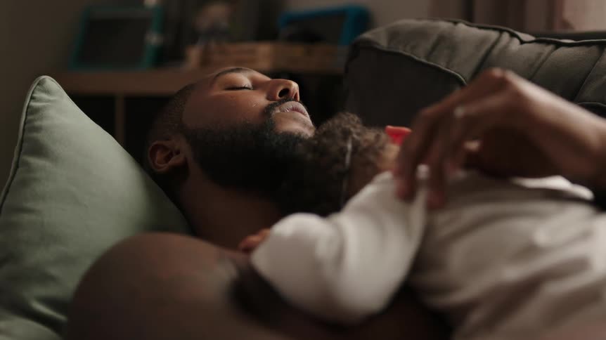 A man in a room laying on a couch holding a baby with a wall in the background