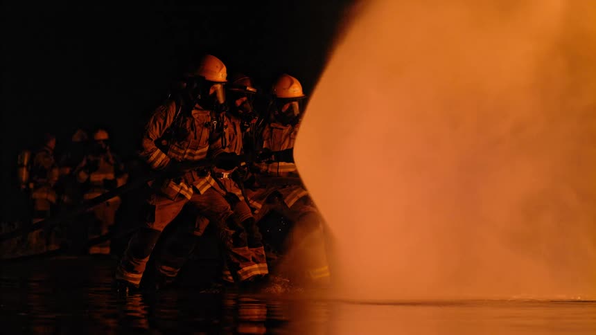 A group of firefighters hold a large fire hose as they spray water on a fire