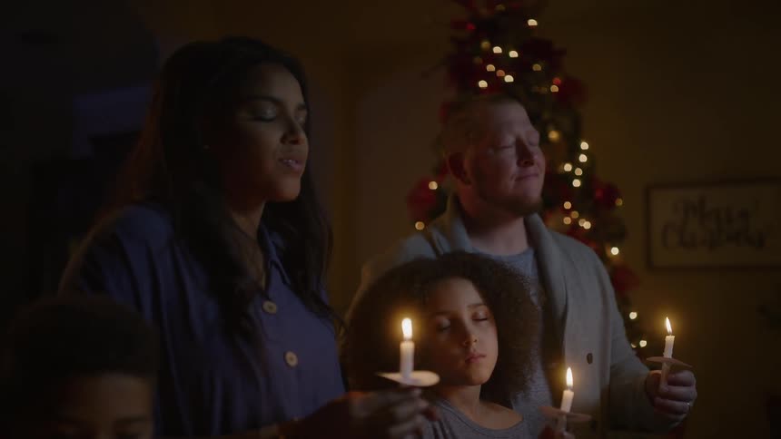 A man and woman glance and smile at one another while holding candles with their children in the dimly lit living room of a house in the evening