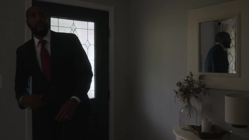 A man walks into a house in a suit and tie and checks himself out in the mirror