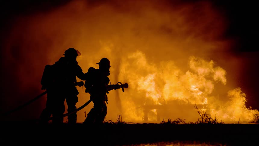 Firefighters holding firehose stand silhouetted by the raging fire outdoors