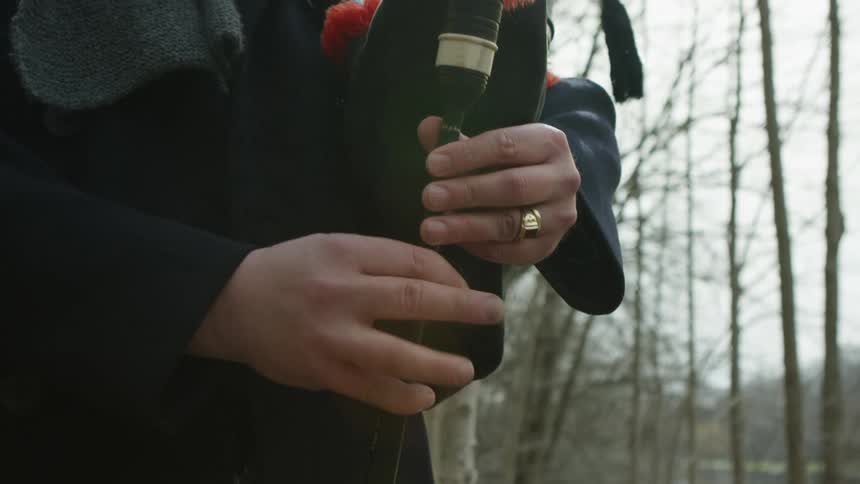 A male bagpipe player stands outdoors near some leafless trees while performing