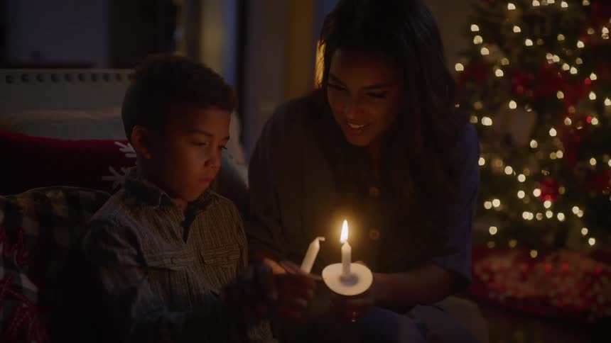 A boy and a woman in a room lighting candles with a christmas tree in the background