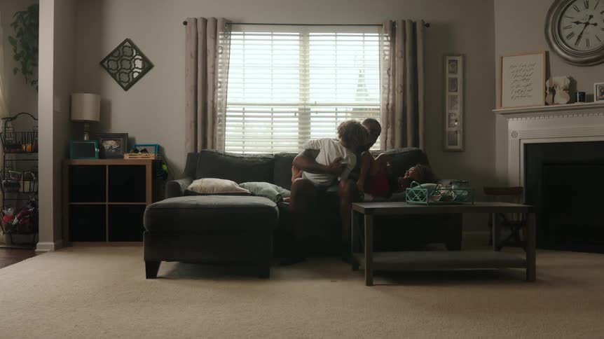 A father playfully wrestles with his two children on a sofa in the daytime