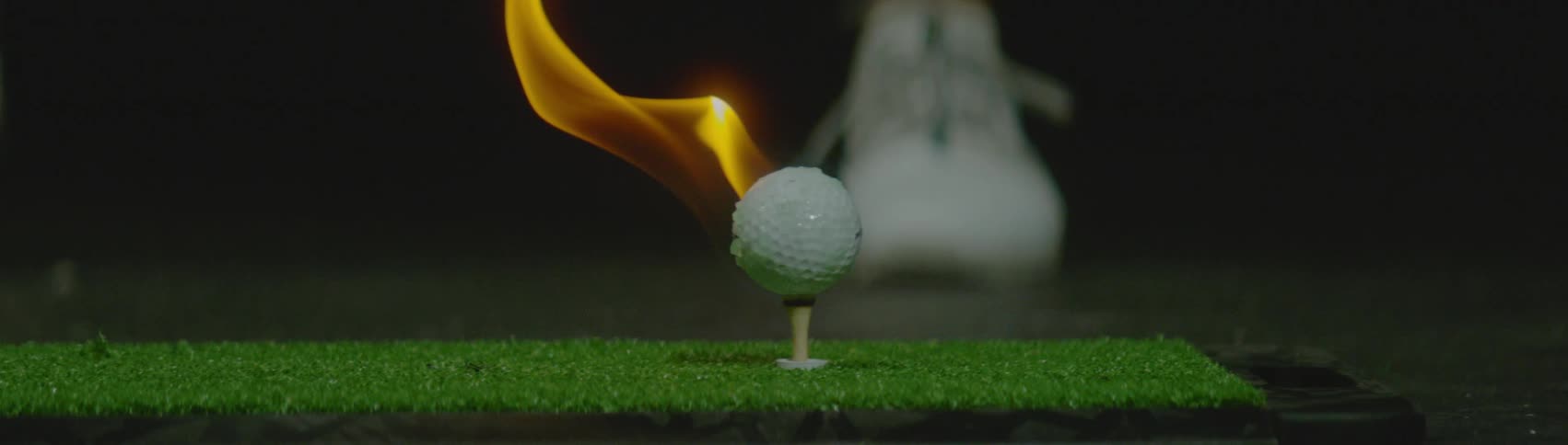A flaming golf ball sits on a tee on green turf before a club strikes it