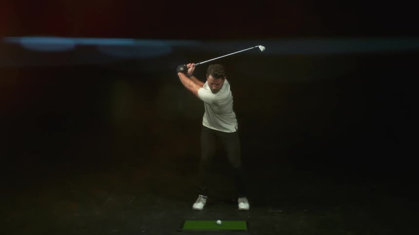 A man swings a golf club and hits a golf ball outside at night