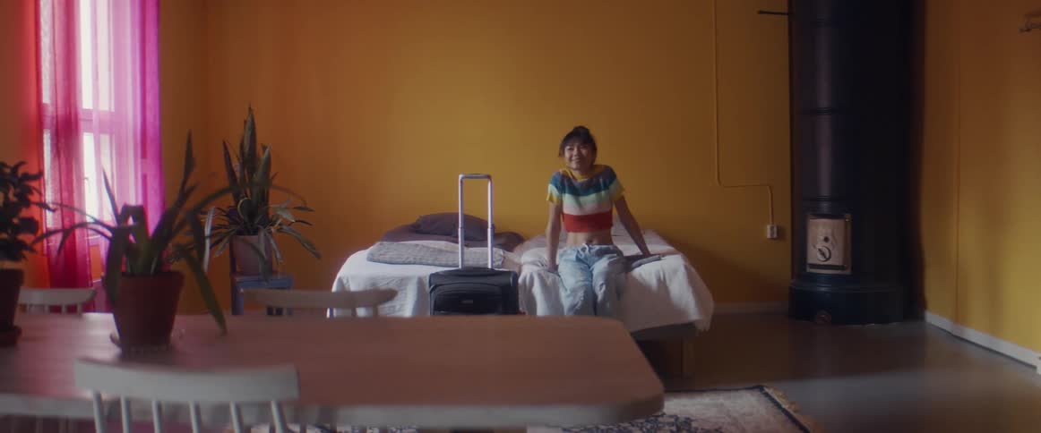 A woman pulls luggage behind her as she approaches a bed to sit for a moment before lying back