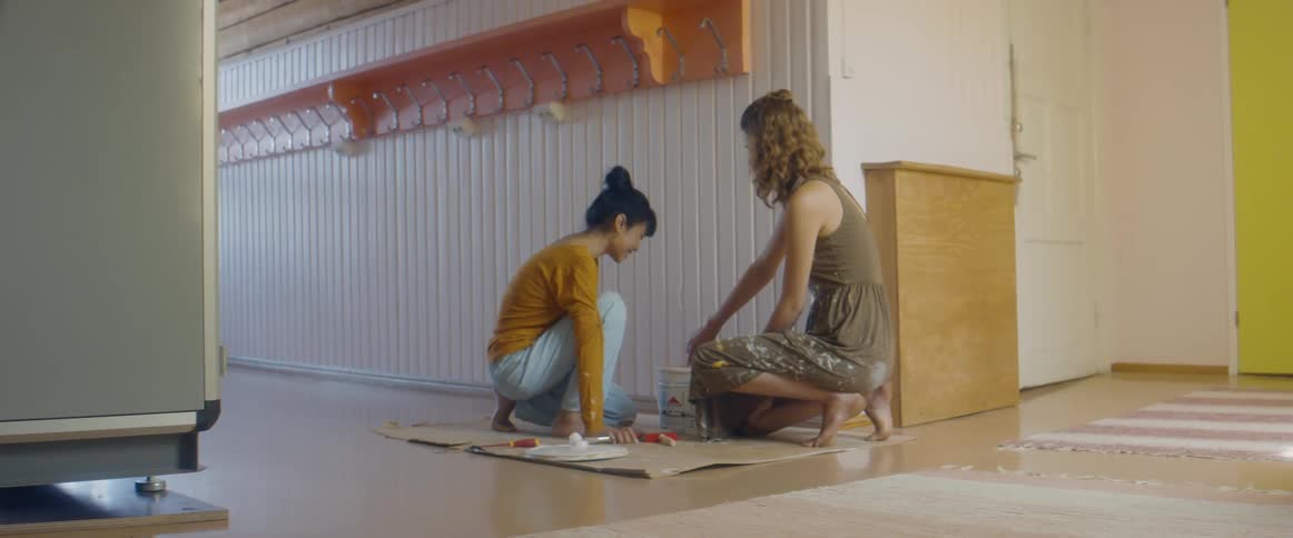 Two women mixing paint on the floor of an empty apartment