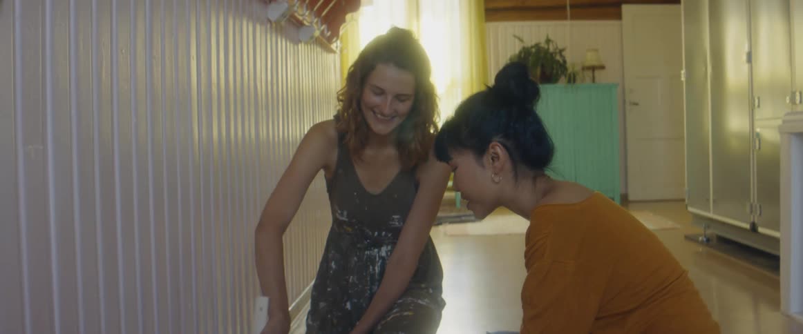 Two women on the floor of a room with one woman mixing paint in a bucket with a wall and window in the background