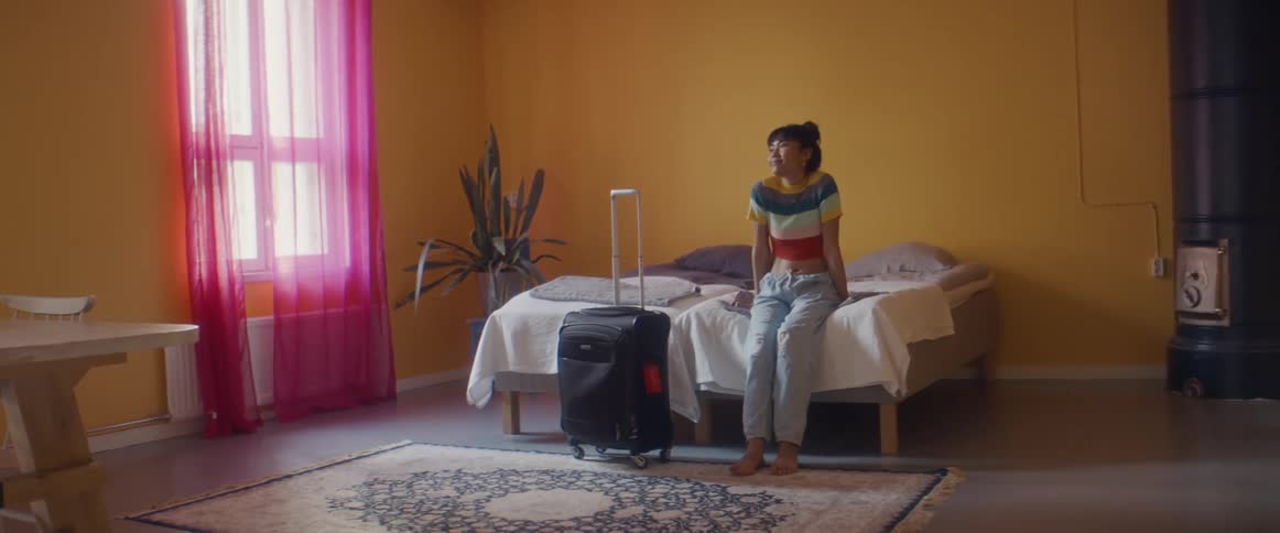 A woman rolls a suitcase into a bedroom and flops on the bed