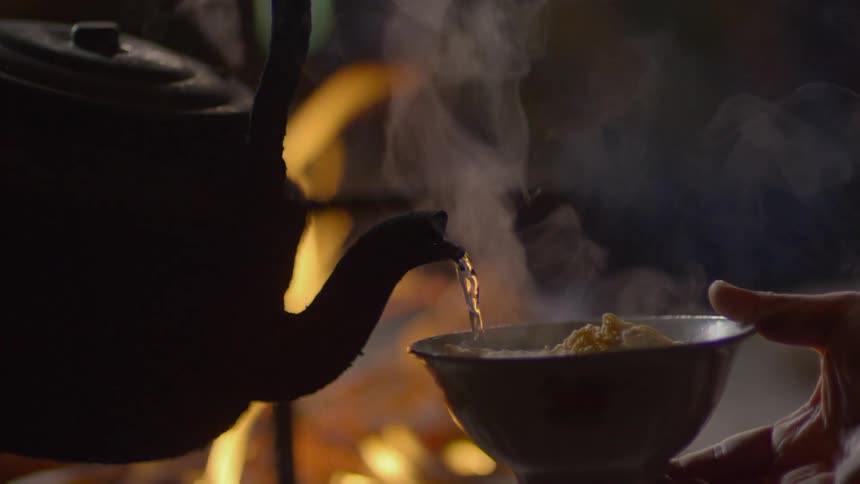 A person holding a bowl and pouring hot water from a kettle into it with fire in the background