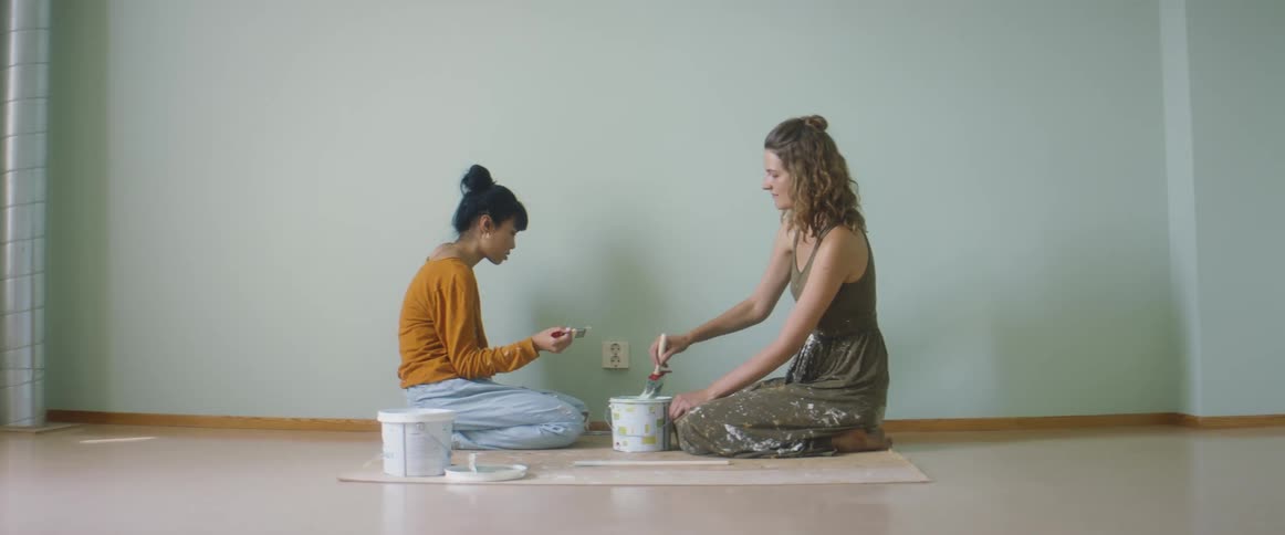 Two women in a room on the floor with paintbrushes next to a can of paint