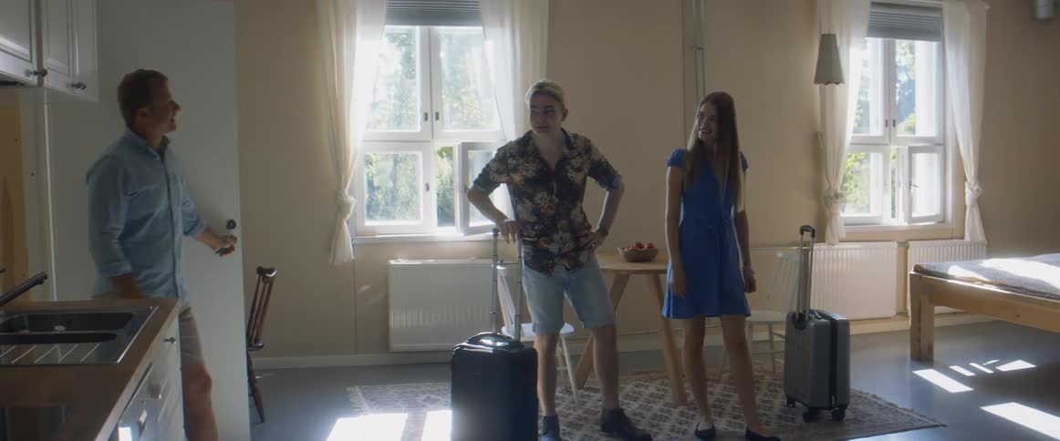 A man and a woman with luggage in a room waving goodbye to another man and a woman with a wall and window in the background