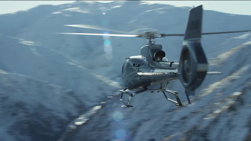 A helicopter hovers in the air by large snow covered mountains
