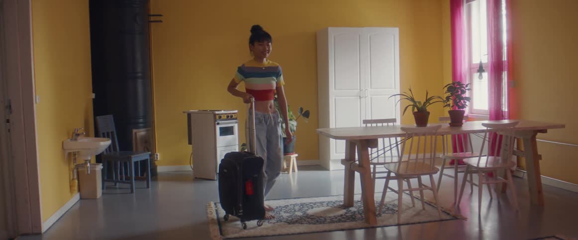 A young woman walks into a small apartment room with her luggage before sitting down