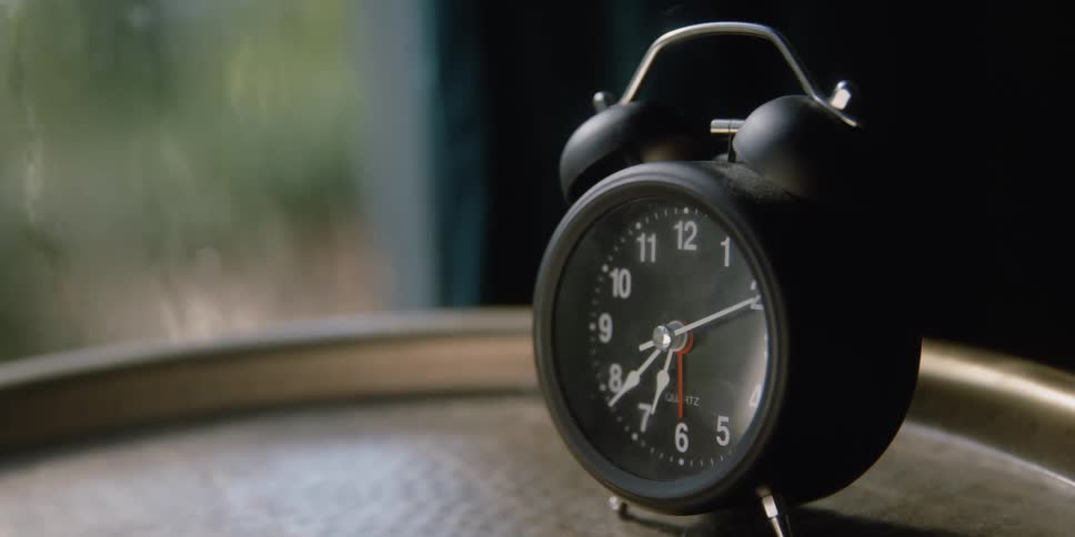 An old timey analog clock sits on a tray in front of a moist window