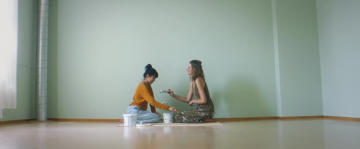 Two women playfully dab at each other with paint brushes near wall painted the same color as that in the paint cans