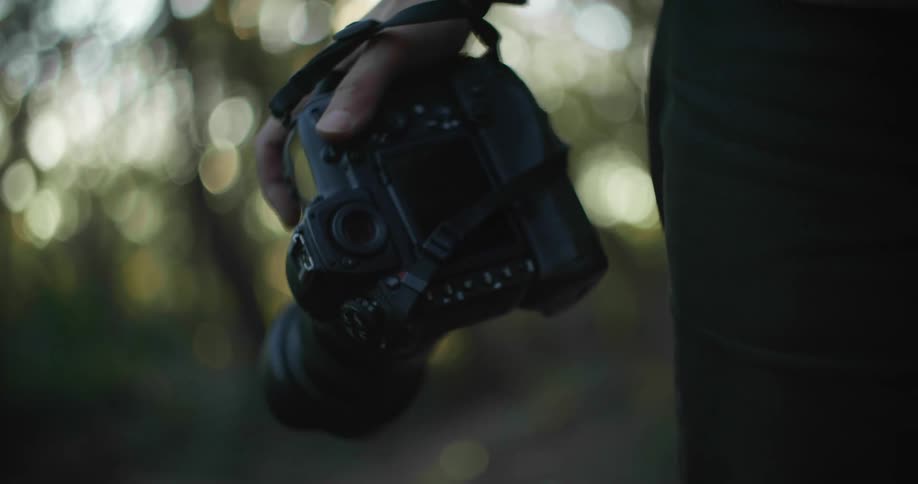A man holds a camera down by his side while walking through the forest