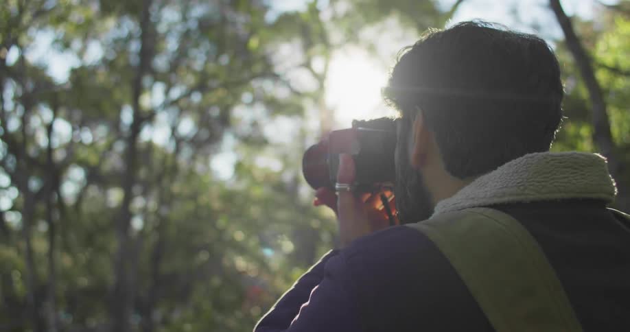A man in a forest with a camera with the sun the sky and clouds in the background