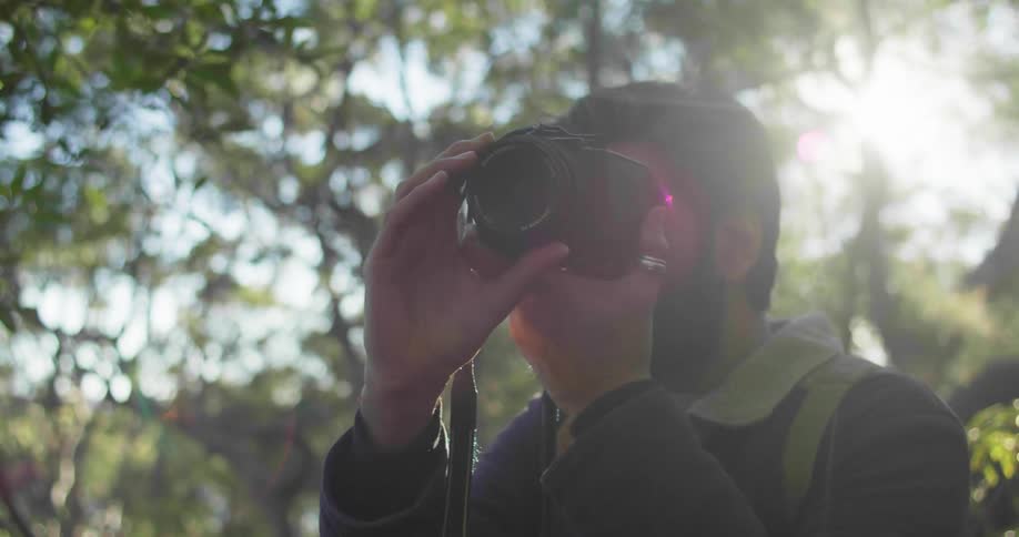 A man with a camera and a backpack in a forest taking a picture with the sky and clouds in the background