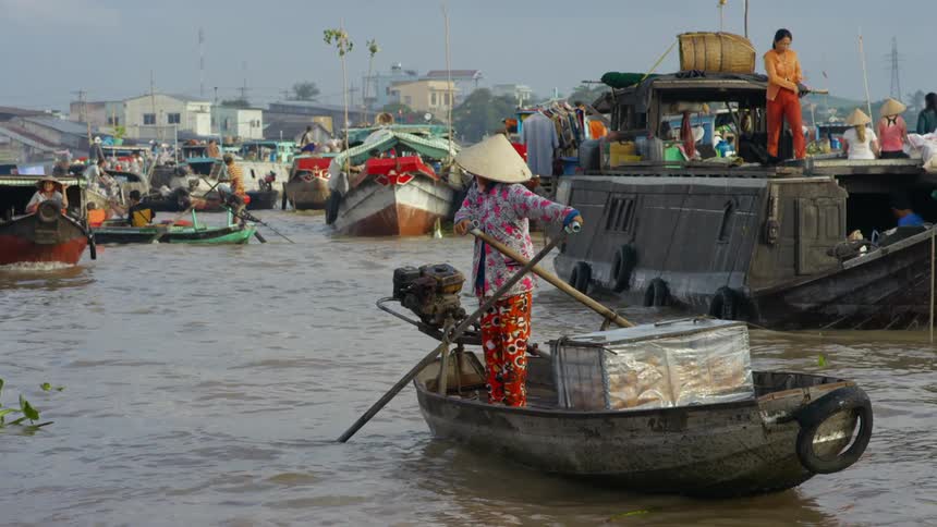 A woman with a hat on a river with a boat with other boats buildings the sky and clouds in the background