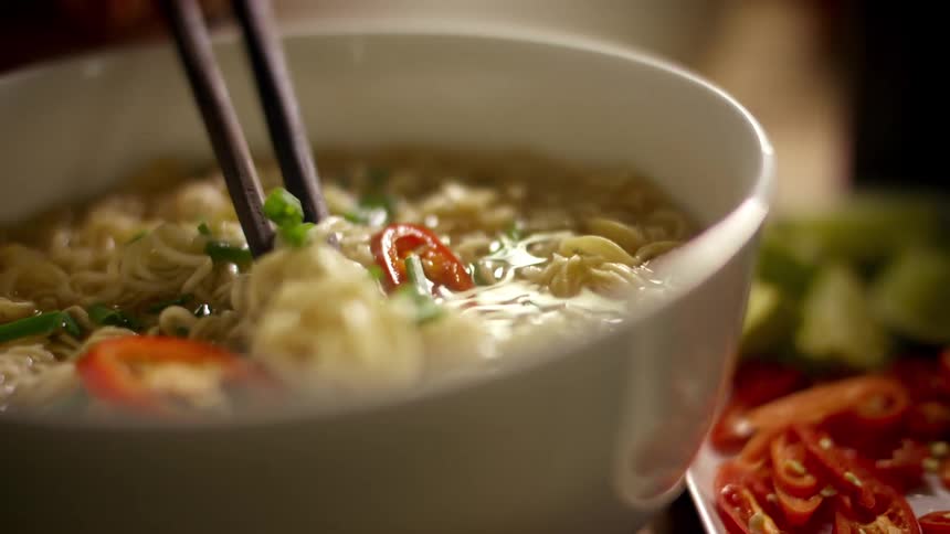 Two people use chopsticks to pick up noodles in a bowl at a restaurant