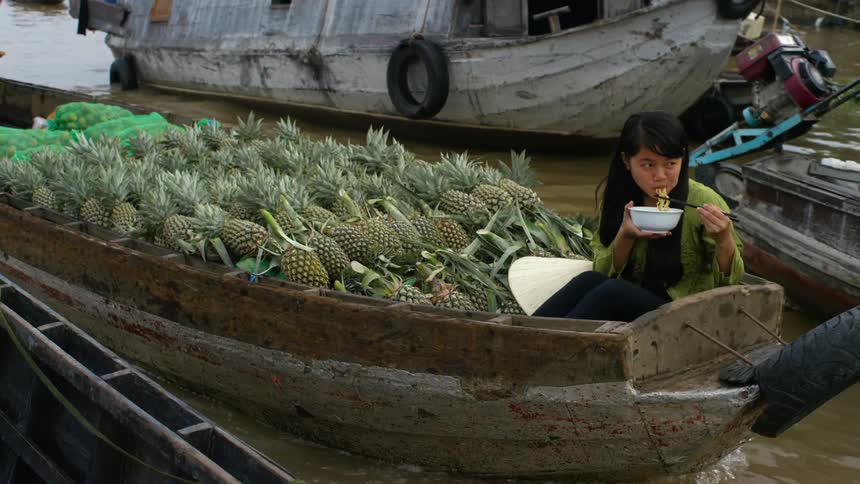 Woman eating a bowl of noodles over a boat loaded with pineapples