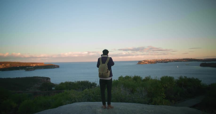 Man wearing backpack stands on black clear stone surface photographing ocean stretching to red tinted horizon