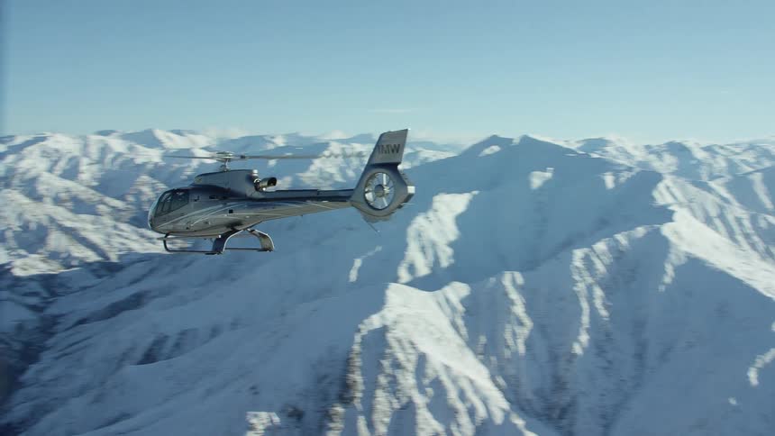 A helicopter flies over a snow capped mountain range on a clear day