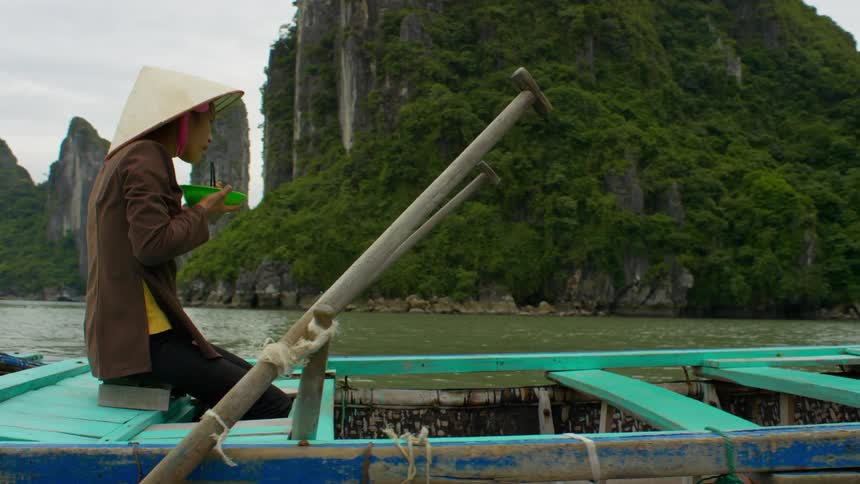 A woman eats from a bowl with chopsticks as she sits in a boat on a river on a cloudy day