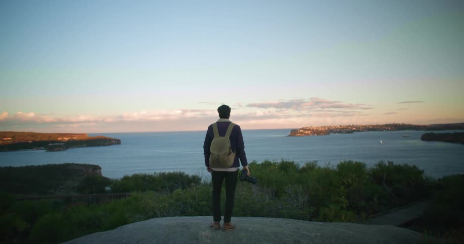A photographer standing on a boulder looks out over a bay in the evening