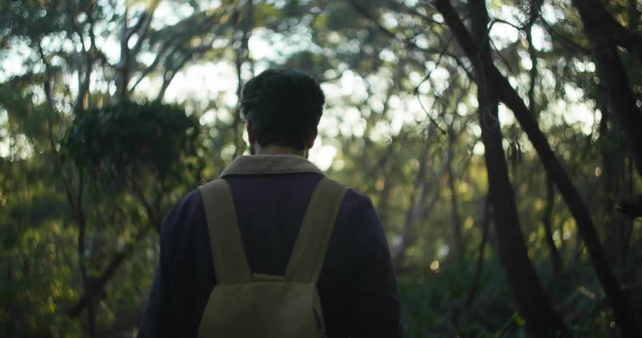 Man wearing jacket and backpack walks through woods looking around