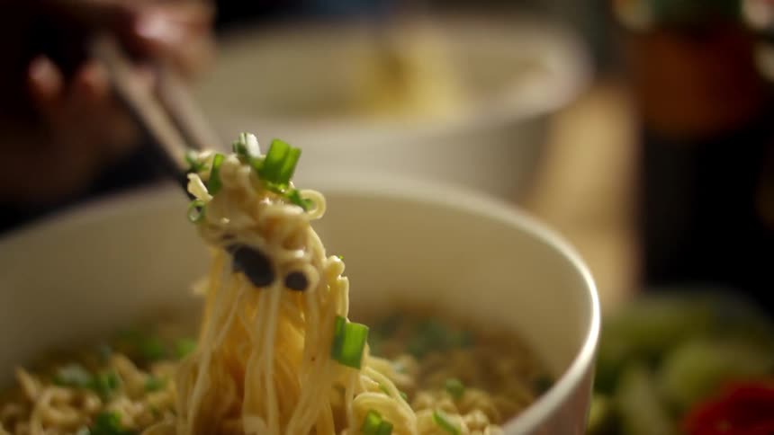 A woman is lifting noodles and green onions with chopsticks at a restaurant table