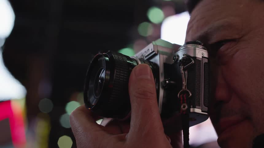 A man in front of lit up buildings takes a photo with a camera at night