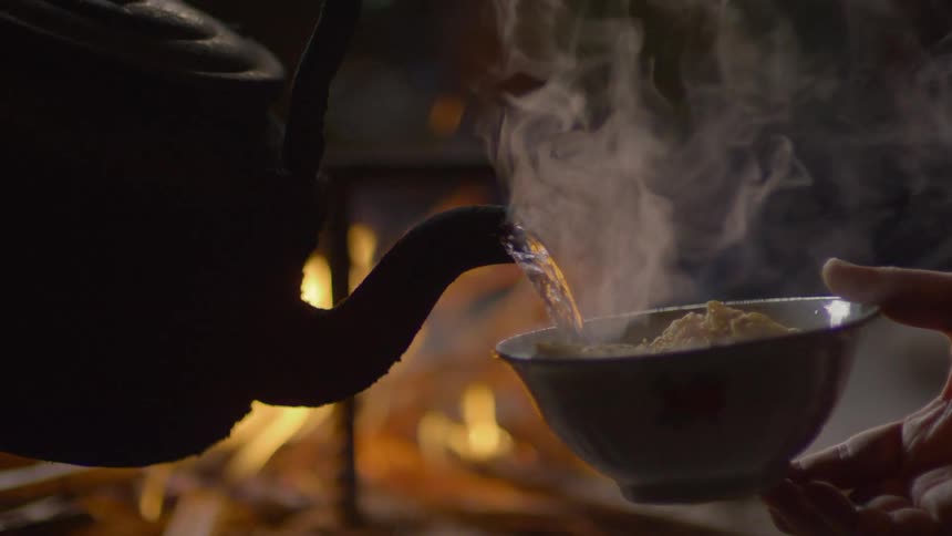 Pouring hot water from an iron teapot into a soup bowl in front of a fire place