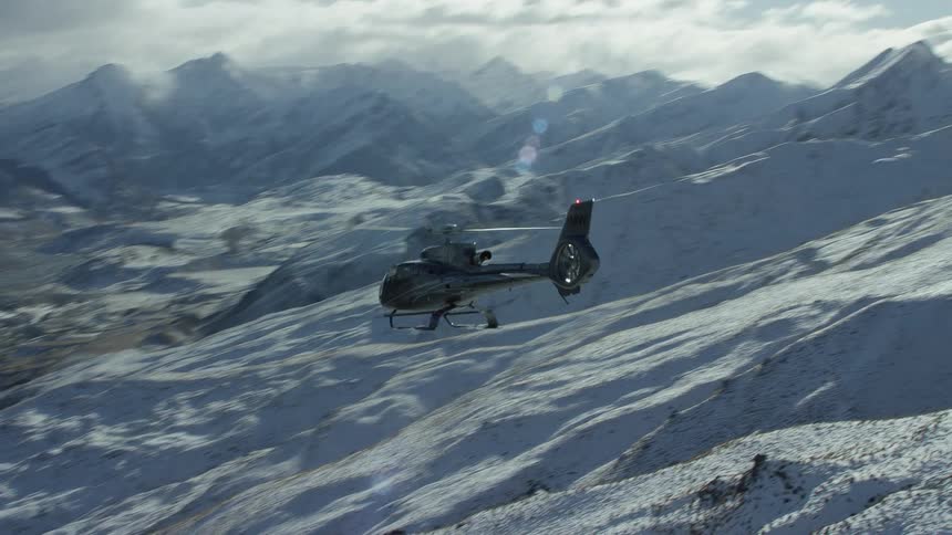 A helicopter hovers over a valley beside snow covered mountains