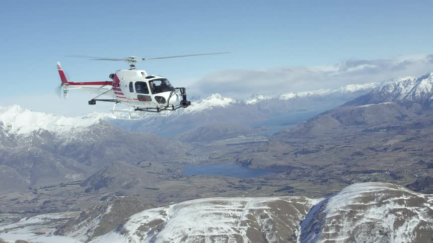 Helicopter moves slowly over snow covered mountain tops with lake occupying center of valley