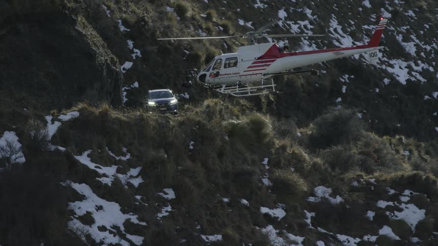 A car drives on a road in a mountain range as a helicopter flies towards the road