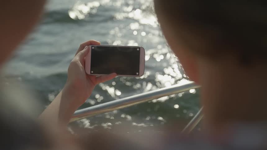 Two people stand on a boat and hold a cell phone over the water