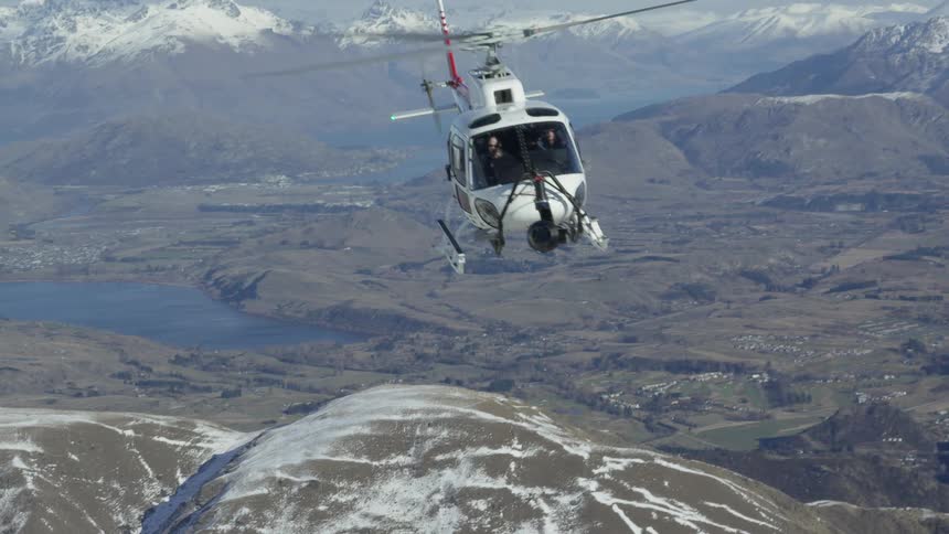 A helicopter hovers over a valley by snow covered mountains
