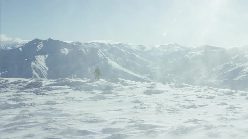 A person is standing on top of a snow covered mountain as the wind blows snow into the air