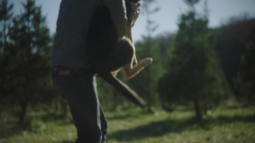 Man wearing knit cap stands holding squirming dog near conifer trees