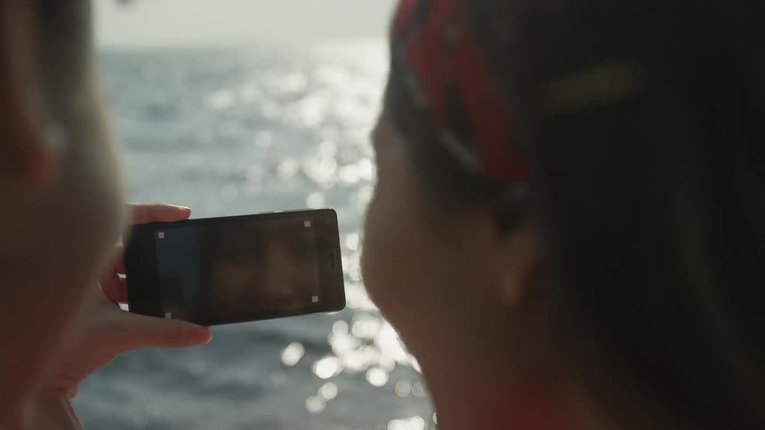 Woman stands near friends laughing as she films ocean waves