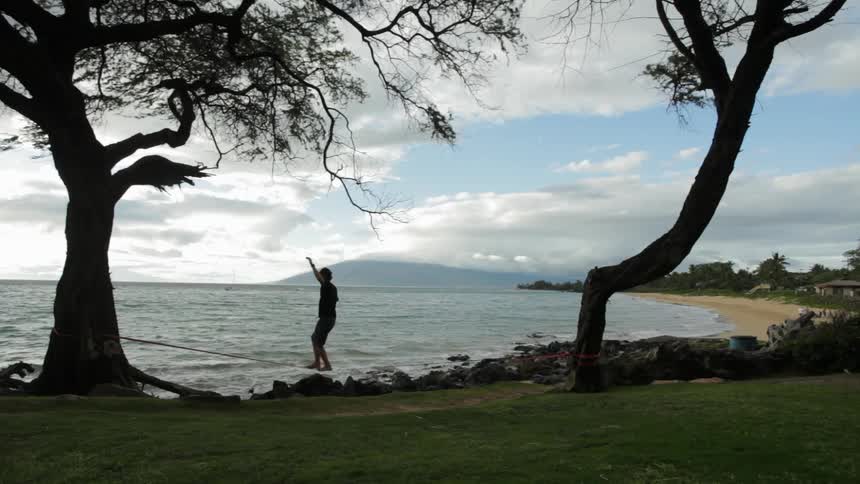 Person practices walking on rope tied between trees near the water