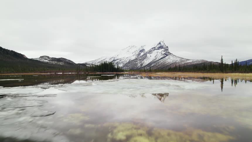 The smooth calm surface of a lake reflects the snow capped mountain and cloudy sky